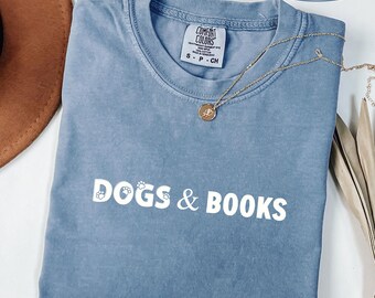 May include: A blue jean colored t-shirt with a crew neckline and short sleeves. The shirt features the white text DOGS & BOOKS printed across the chest, with small paw prints integrated into the letters. A gold chain necklace rests on the fabric.