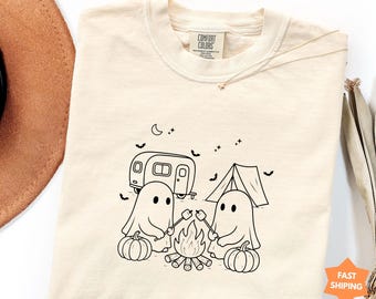 Può includere: Una t-shirt a maniche corte color avorio con un disegno a linee nere di due fantasmi che arrostiscono marshmallow su un falò. La scena include un camper, una tenda, zucche, pipistrelli e una luna crescente. L'etichetta interna riporta Comfort Colors.