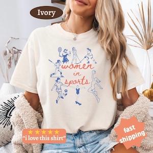 May include: Cream-coloured t-shirt featuring a blue and red graphic of women in sports. The words "women in sports" are displayed. The shirt also has the text "I love this shirt" and "fast shipping".