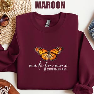 May include: Maroon sweatshirt featuring a vibrant monarch butterfly design. The butterfly is orange, black, and white. The text "made for more" and "Ephesians 2:10" are printed in white script below the butterfly. The sweatshirt is a solid maroon color.