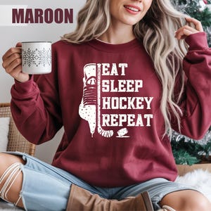 Könnte beinhalten: Burgunderfarbenes Sweatshirt mit dem weißen Text "EAT SLEEP HOCKEY REPEAT" und einem Hockey-Schlittschuh-Motiv. Eine weiße Tasse mit Schneeflockenmuster wird gehalten. Das Wort "MAROON" steht oben.