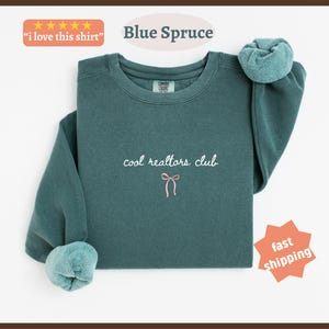 May include: A blue spruce colored crewneck sweatshirt featuring the embroidered white text "cool realtors club" centered on the chest, positioned above a small, simple pink line-art bow graphic. The garment has long sleeves with ribbed cuffs.