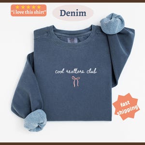 May include: A denim blue crewneck sweatshirt featuring the embroidered white text "cool realtors club" centered on the chest, positioned above a small, simple pink bow graphic. The garment has long sleeves with ribbed cuffs and a soft interior lining.
