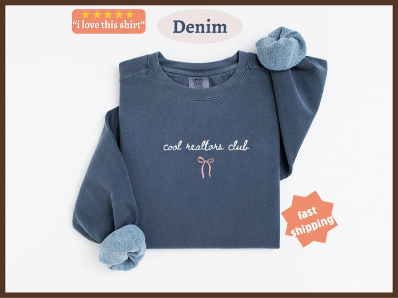 May include: A denim blue crewneck sweatshirt featuring the embroidered white text "cool realtors club" centered on the chest, positioned above a small, simple pink bow graphic. The garment has long sleeves with ribbed cuffs and a soft interior lining.