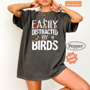 May include: A charcoal gray short-sleeve t-shirt featuring the white text "EASILY DISTRACTED BY BIRDS" and colorful illustrations of four different birds perched on branches. The shirt has a crew neckline and a relaxed, loose-fitting silhouette.