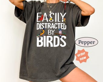 Może przedstawiać: Ciemnoszary t-shirt z krótkim rękawem z białym napisem "EASILY DISTRACTED BY BIRDS" oraz kolorowymi ilustracjami czterech różnych ptaków siedzących na gałęziach. Koszulka ma okrągły dekolt i luźny, swobodny krój.