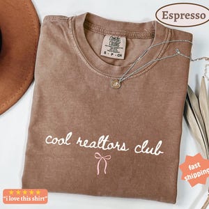 May include: A brown short-sleeve t-shirt with a crew neckline and a Comfort Colors brand label. The front features the white text "cool realtors club" in a cursive font, positioned above a small, simple pink line-drawn bow graphic.