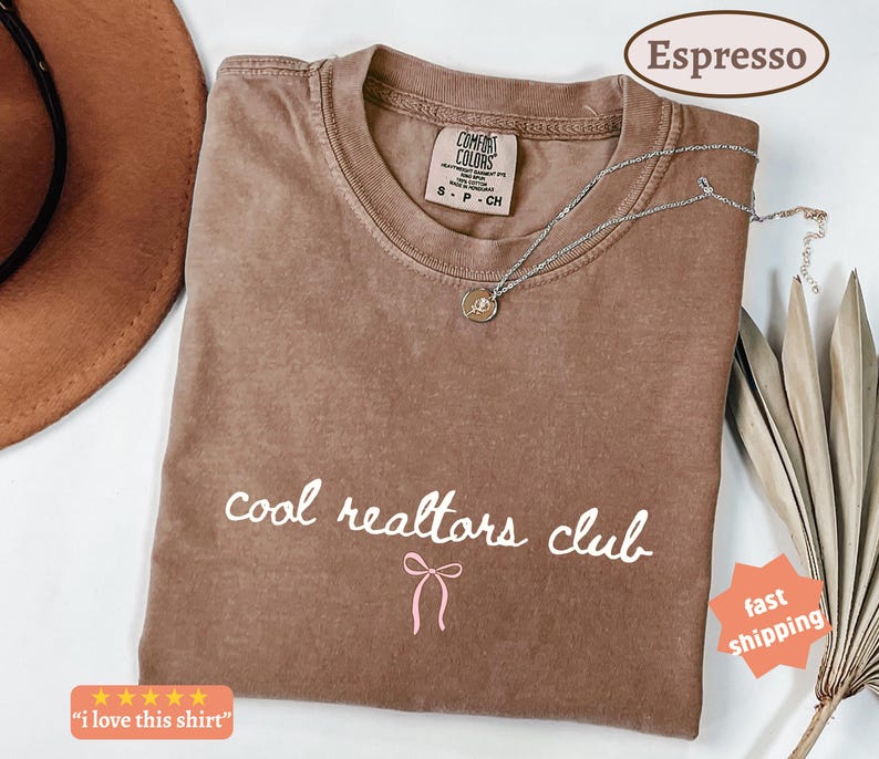 May include: A brown short-sleeve t-shirt with a crew neckline and a Comfort Colors brand label. The front features the white text "cool realtors club" in a cursive font, positioned above a small, simple pink line-drawn bow graphic.