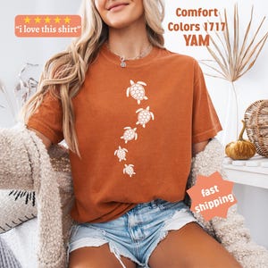 May include: Burnt orange t-shirt with a vertical line of white sea turtle illustrations. The shirt has the text "I love this shirt" in a small orange box. The shirt is part of the "Comfort Colors 1717 YAM" collection.