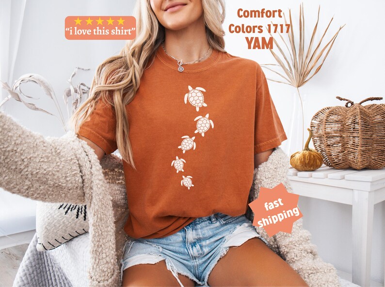 May include: Burnt orange t-shirt with a vertical line of white sea turtle illustrations. The shirt has the text "I love this shirt" in a small orange box. The shirt is part of the "Comfort Colors 1717 YAM" collection.
