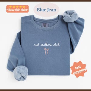 May include: A blue jean colored crewneck sweatshirt featuring the embroidered white text "cool realtors club" centered on the chest, positioned above a small, simple pink bow graphic. The garment has long sleeves with gathered cuffs.