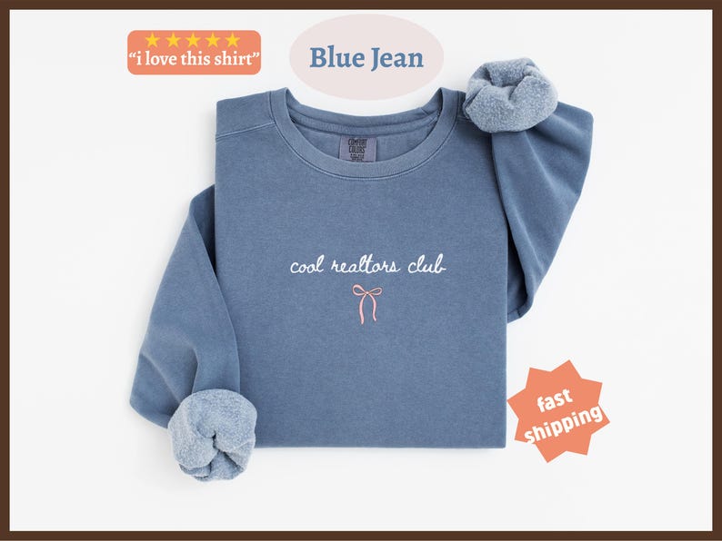May include: A blue jean colored crewneck sweatshirt featuring the embroidered white text "cool realtors club" centered on the chest, positioned above a small, simple pink bow graphic. The garment has long sleeves with gathered cuffs.
