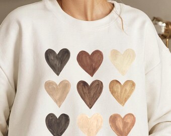 Pode incluir: Camisola branca com nove corações de diferentes cores impressos na frente. Os corações estão dispostos em três filas de três. As cores dos corações são castanho, bege e castanho escuro.