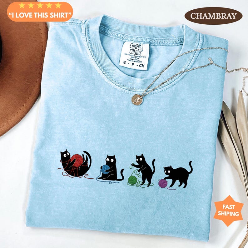 May include: Light blue Comfort Colors t-shirt featuring a playful design of four black cats interacting with yarn balls in red, blue, green, and purple. The shirt has the text "I LOVE THIS SHIRT" and "CHAMBRAY".