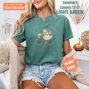 May include: Light green t-shirt featuring a frog on a bicycle graphic. The frog is dressed in a dress and scarf, with a basket of flowers on the bike. The shirt has the text "off to cause a kerfuffle at mahjong" and "Comfort Colors 1717 LIGHT GREEN".
