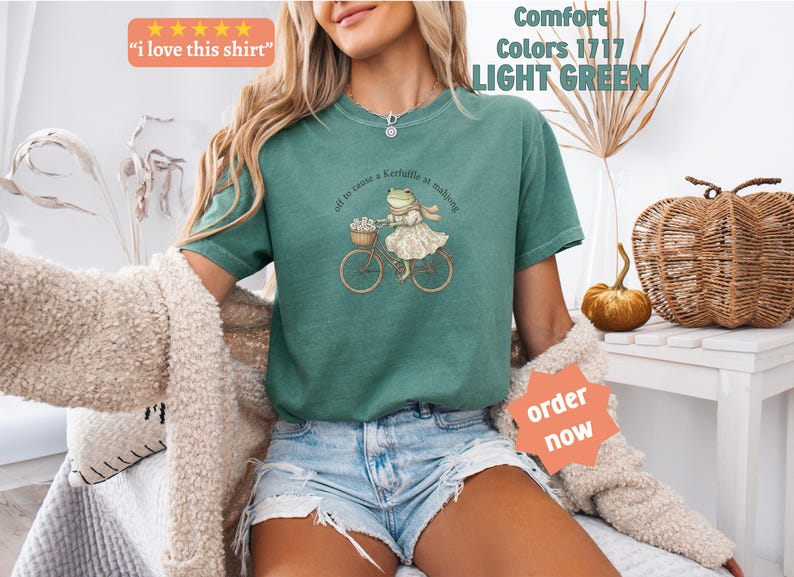May include: Light green t-shirt featuring a frog on a bicycle graphic. The frog is dressed in a dress and scarf, with a basket of flowers on the bike. The shirt has the text "off to cause a kerfuffle at mahjong" and "Comfort Colors 1717 LIGHT GREEN".