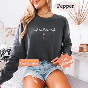 May include: A charcoal gray long-sleeve sweatshirt featuring the white text "cool realtors club" and a small pink bow graphic centered on the chest. The garment has a crew neckline and long sleeves, paired with light blue denim shorts.