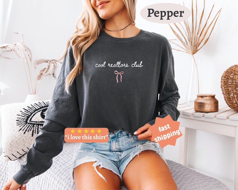 May include: A charcoal gray long-sleeve sweatshirt featuring the white text "cool realtors club" and a small pink bow graphic centered on the chest. The garment has a crew neckline and long sleeves, paired with light blue denim shorts.