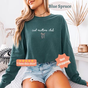 May include: A dark teal long-sleeve crewneck sweatshirt featuring the white text "cool realtors club" in a cursive font, centered above a small, simple pink line-art bow graphic. The garment has a relaxed fit and a solid, muted color.