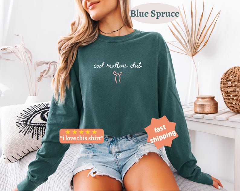 May include: A dark teal long-sleeve crewneck sweatshirt featuring the white text "cool realtors club" in a cursive font, centered above a small, simple pink line-art bow graphic. The garment has a relaxed fit and a solid, muted color.