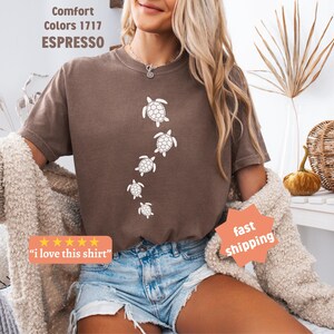 May include: Espresso-colored t-shirt featuring a white turtle design. The shirt has a crew neck and short sleeves. The text "Comfort Colors 1717 ESPRESSO" is visible. The shirt is paired with denim shorts and a cream-colored cardigan.