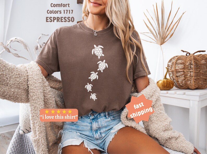May include: Espresso-colored t-shirt featuring a white turtle design. The shirt has a crew neck and short sleeves. The text "Comfort Colors 1717 ESPRESSO" is visible. The shirt is paired with denim shorts and a cream-colored cardigan.