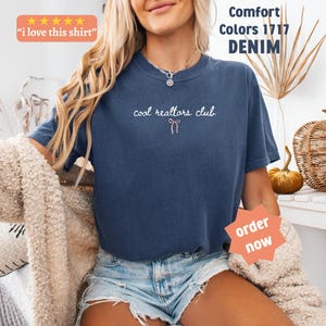 May include: A denim blue short-sleeve t-shirt featuring the white text "cool realtors club" with a small pink bow graphic centered below the text. The shirt includes a crew neckline and is labeled as Comfort Colors 1717 in the color denim.