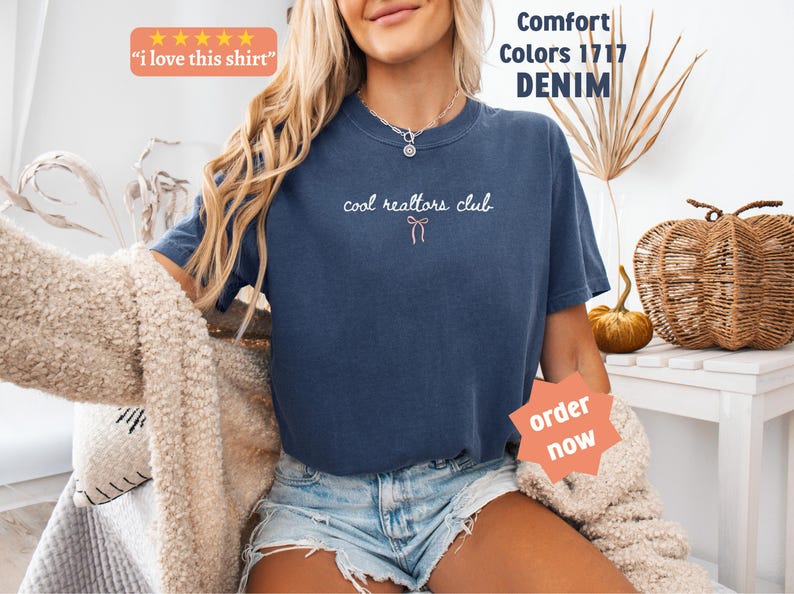 May include: A denim blue short-sleeve t-shirt featuring the white text "cool realtors club" with a small pink bow graphic centered below the text. The shirt includes a crew neckline and is labeled as Comfort Colors 1717 in the color denim.