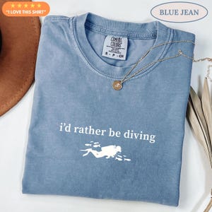 Könnte beinhalten: Ein blaues T-Shirt mit dem Text "i'd rather be diving" und einer Tauchergrafik. Das Shirt hat einen Rundhalsausschnitt und kurze Ärmel. Eine goldene Halskette mit Anhänger ist auf dem Shirt drapiert. Das Shirt ist mit "Blue Jean" beschriftet.