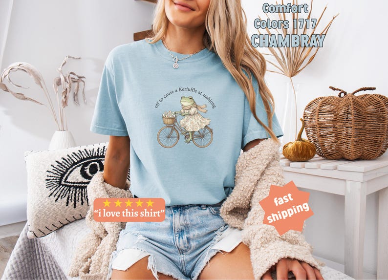 May include: Pale blue t-shirt featuring a frog on a bicycle, with the text "off to cause a Kerfuffle at mahjong". The shirt also has the text "Comfort Colors 1717 CHAMBRAY".