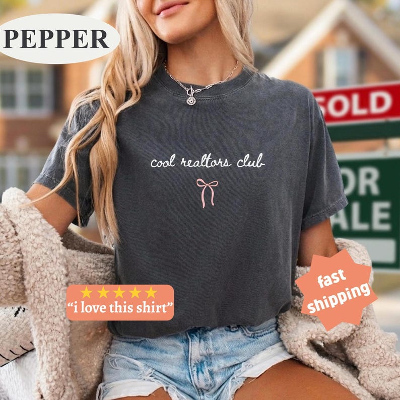 May include: A charcoal gray short-sleeve t-shirt featuring the white text "cool realtors club" printed on the chest, accompanied by a small, simple pink line-art bow graphic centered directly below the text.