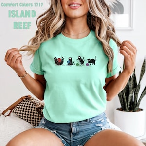 May include: A light green t-shirt with the text "ISLAND REEF" and a graphic of four black cats playing with yarn balls. The shirt is made of Comfort Colors 1717 material.