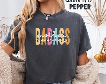 May include: Dark gray t-shirt with the words "BADASS mother runner" in a colorful, retro font. The shirt also has the words "Comfort Colors 1717 PEPPER" printed on the top right corner.