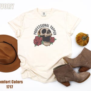 Puede incluir: Camiseta de color marfil con un gráfico de calavera, rosas y el texto "PROFESSIONAL YAPPER". La imagen incluye un sombrero marrón, botas marrones y una pequeña calabaza. La camiseta es Comfort Colors 1717.