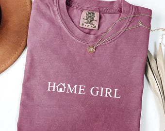 May include: Berry-colored t-shirt with the text "HOME GIRL" in white. The shirt has a crew neck and short sleeves. A gold necklace with a pendant is draped across the shirt. The shirt is labeled "Comfort Colors".