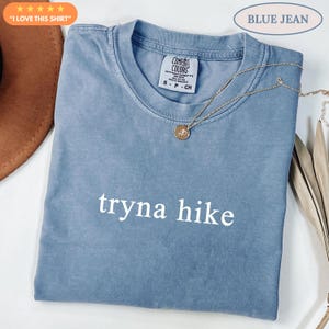 May include: A blue t-shirt with the text "tryna hike" in white. The shirt has a crew neck and short sleeves. A gold necklace with a pendant is draped across the shirt. The text "BLUE JEAN" is above the shirt.