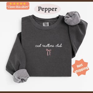 May include: A charcoal gray crewneck sweatshirt featuring the embroidered white text "cool realtors club" centered on the chest, positioned above a small, simple pink embroidered bow graphic. The garment has long sleeves with ribbed cuffs.