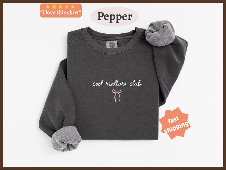 May include: A charcoal gray crewneck sweatshirt featuring the embroidered white text "cool realtors club" centered on the chest, positioned above a small, simple pink embroidered bow graphic. The garment has long sleeves with ribbed cuffs.