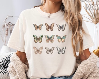 Peut inclure: T-shirt de couleur ivoire avec neuf illustrations détaillées de papillons de différentes couleurs. Les papillons sont disposés en trois rangées de trois. Le t-shirt a une coupe décontractée et des manches courtes. Le texte "Comfort Colors 17" et "IVORY" est visible en haut à droite.