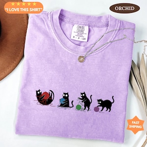 May include: A light purple t-shirt with a crew neck and short sleeves. The shirt features a graphic of four black cats playing with yarn in red, blue, green, and purple. The text "I LOVE THIS SHIRT" is in the upper left corner.