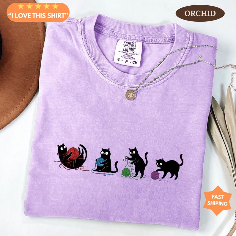 May include: A light purple t-shirt with a crew neck and short sleeves. The shirt features a graphic of four black cats playing with yarn in red, blue, green, and purple. The text "I LOVE THIS SHIRT" is in the upper left corner.