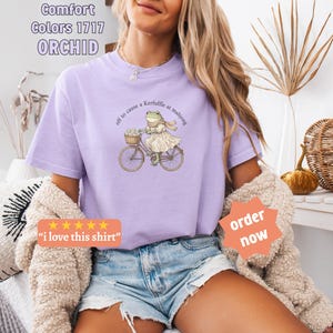 May include: A lilac t-shirt featuring a frog on a bicycle, with the phrase "off to cause a Kerfuffle at mahjong." The shirt also has the text "Comfort Colors 1717 ORCHID". The shirt is a light purple colour.