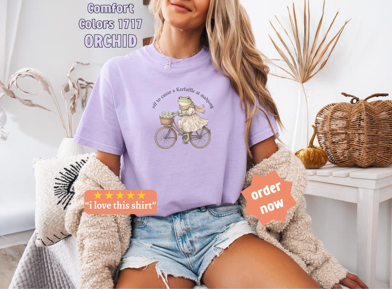 May include: A lilac t-shirt featuring a frog on a bicycle, with the phrase "off to cause a Kerfuffle at mahjong." The shirt also has the text "Comfort Colors 1717 ORCHID". The shirt is a light purple colour.