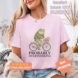 May include: Pale pink t-shirt featuring a frog on a bicycle with a cat in the basket, and the words "PROBABLY OVERTHINKING". The shirt also has the text "Comfort Colors 1717 BLOSSOM".
