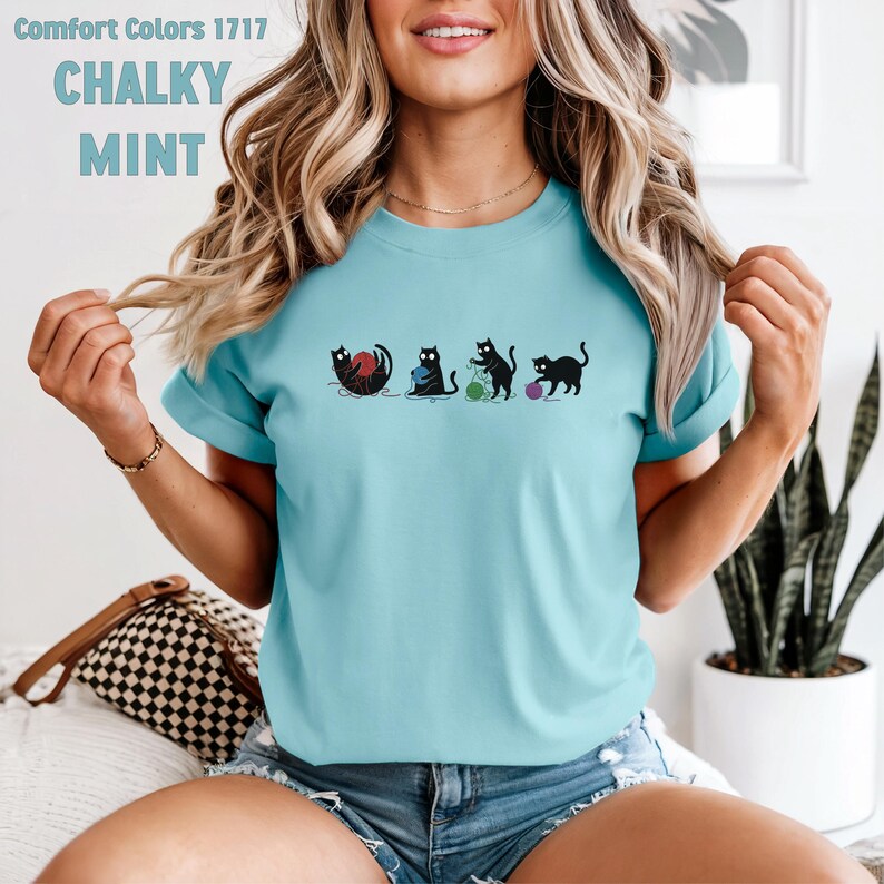 May include: A light blue t-shirt with the words "Comfort Colors 1717" and "CHALKY MINT" printed at the top. The shirt features a graphic of four black cats playing with yarn. The cats are in different poses, with yarn balls in red, green, and purple.
