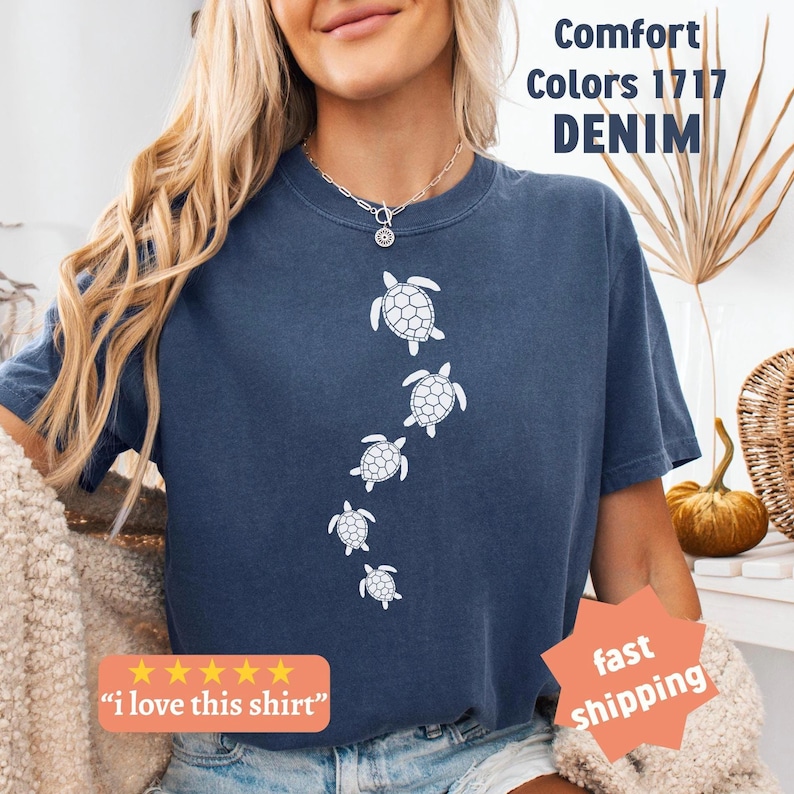 May include: A denim blue t-shirt featuring a series of white sea turtle illustrations. The shirt has the text "Comfort Colors 1717 DENIM" and a five-star rating with the text "i love this shirt".