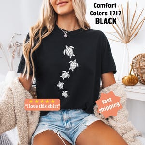 May include: Black t-shirt featuring a series of white sea turtle illustrations. The shirt has the text "Comfort Colors 1717 BLACK" and a small orange sticker that says "i love this shirt".