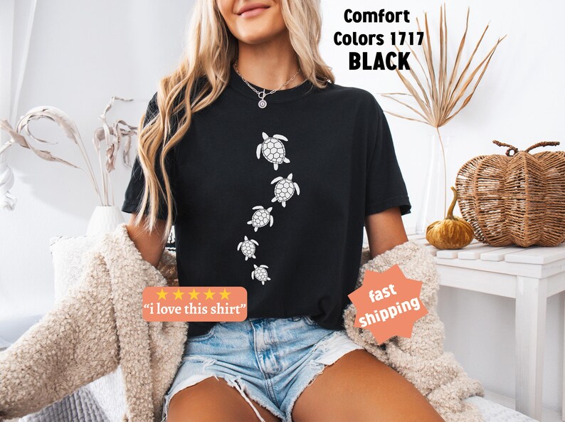 May include: Black t-shirt featuring a series of white sea turtle illustrations. The shirt has the text "Comfort Colors 1717 BLACK" and a small orange sticker that says "i love this shirt".
