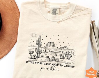 May include: An ivory short-sleeve t-shirt featuring a black line-art desert landscape with cacti, mountains, and a starry sky. The text reads, "IF THE STARS WERE MADE TO WORSHIP so will i." The shirt has a Comfort Colors brand label at the inner neck.