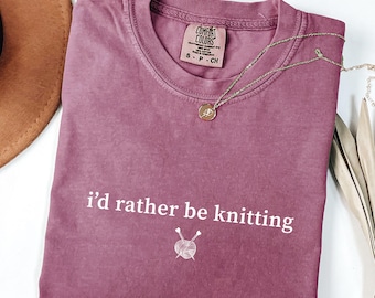 May include: A berry-colored short-sleeve t-shirt with a crew neckline. The shirt features the white text 'i'd rather be knitting' above a small graphic of a ball of yarn with two knitting needles. A gold-toned necklace rests on the shirt.